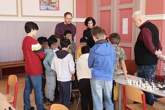 Eine Gruppe Kinder und drei Erwachsene stehen um einen Tisch an dem Schach gespielt wird. 