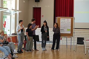 Fünf Menschen vor einer beschriebenen Stelltafel stehend und diskutierend.  Im Raum sitzen weitere Menschen im Halbkreis angedeutet. Im Hintergrund des Raumes eine Bühne mit Vorhang und einer Leinwand. 