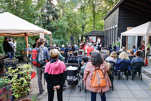 Publikum sitzt im Freien vor einer kleinen Bühne und lauscht einem Musikauftritt beim Kulturfestival „Salon K“ im Lettekiez.