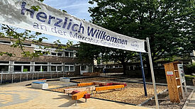 Eine Außenfläche mit Balancierstäben, einer Bücherbox, Sitzbänken und Bäumen. Im Vordergrund sieht man ein Banner mit der Aufschrift „Herzlich Willkommen an der Mercator-Grundschule“. 