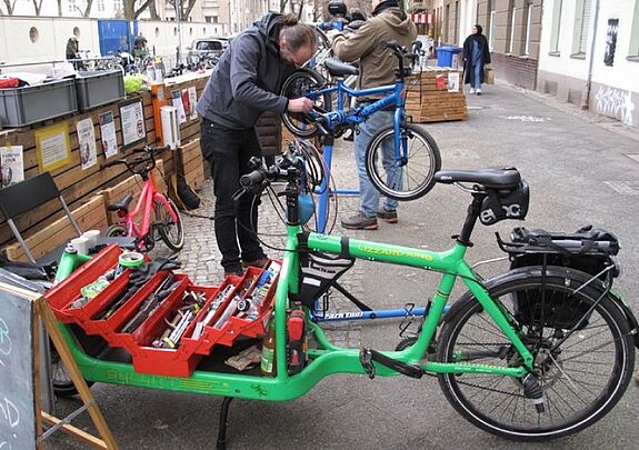 Ein Mechaniker repariert ein Fahrrad, während Werkzeug in einem Lastenrad bereitliegt.