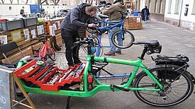 Ein Mechaniker repariert ein Fahrrad, während Werkzeug in einem Lastenrad bereitliegt.