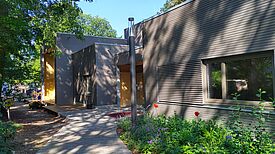 Blick über einen gepflasterten Zugangsweg hin zum Eingangsbereich des Neubaus des Moabiter Kinderhofs. Sonnenstrahlen fallen durch die Bäume im Umfeld auf die klaren kubischen Elemente des Baus mit seiner einheitlichen Fassade.