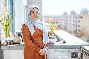 Eine weibliche Person auf einem Balkon eines Wohnhochhauses, sie trägt ein orange-braunes Kleid und ein hellblaues Kopftuch, sie lächelt in die Kamera. Im Hintergrund auf dem Balkon Pflanzen und Töpfe und ferner Wohnungsneubauten. 