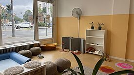 In einem hellen Raum der Kita Am Nettelbeckplatz befinden sich Sitzkissen, ein Bücherregal und Spielmaterialien. Eine große Fensterfront zeigt den Blick auf eine belebte Straße in Berlin-Wedding.