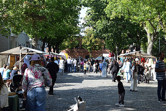 Straßenfest mit Ständen und vielen Menschen unter Bäumen, die gemeinsam Musik, Essen und Spiele genießen.