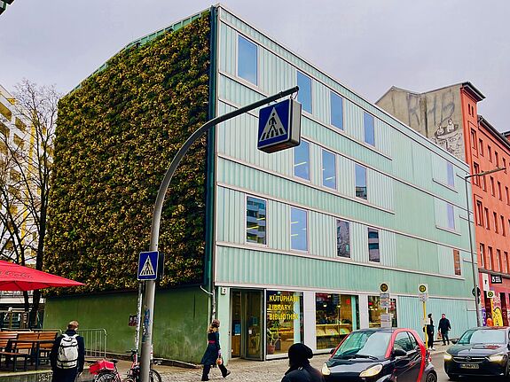 Ein Neubau mit Schaufenstern im Erdgeschoss und begrünter Fassade angrenzend an ein rötliches Wohnhaus. Davor Passanten auf dem Fußweg und parkende Autos an deiner Straße.