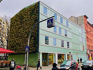 Ein Neubau mit Schaufenstern im Erdgeschoss und begrünter Fassade angrenzend an ein rötliches Wohnhaus. Davor Passanten auf dem Fußweg und parkende Autos an deiner Straße.