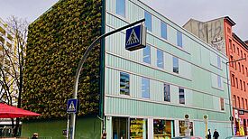 Ein Neubau mit Schaufenstern im Erdgeschoss und begrünter Fassade angrenzend an ein rötliches Wohnhaus. Davor Passanten auf dem Fußweg und parkende Autos an deiner Straße.