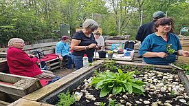 Ein Gruppe von Menschen stehen und sitzen in einem Garten mit Hochbeeten. Sie arbeiten an den Pflanzen und unterhalten sich.  