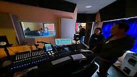Eine Person steht im Aufnahmeraum des Tonstudios und singt in ein Mikrofon, während eine andere Person vor dem Mischpult steht und den Ton überwacht.