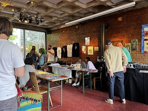 Menschen tauschen sich an Tischen mit Kunstwerken und handgemachten Produkten in der Kugelbar aus; an der Wand hängen Bilder und T-Shirts, im Hintergrund sorgt ein DJ für Musik.