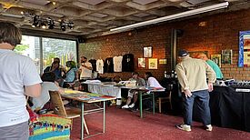 Menschen tauschen sich an Tischen mit Kunstwerken und handgemachten Produkten in der Kugelbar aus; an der Wand hängen Bilder und T-Shirts, im Hintergrund sorgt ein DJ für Musik.