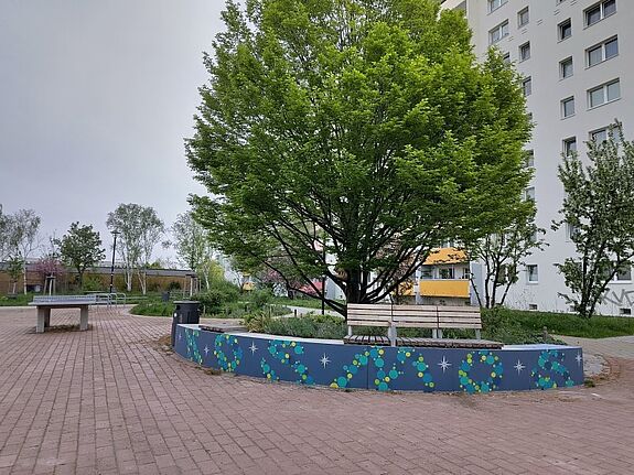 Eine bemalte Betonbank mit bunten Kreisen und Sternen steht unter einem Baum auf dem Satellitenplatz, daneben eine Sitzbank.