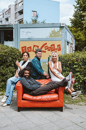 uf einem roten Sofa, das draußen auf einem Weg vor einer Hecke steht, liegen und sitzen vier Menschen in lockerer Pose, sie lächeln. Hinter ihnen ein Funktionsbau mit der Aufschrift „The Corner“ sowie Wohnungsneubauten und Bäume.
