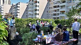 Menschen sitzen und stehen auf einer Grünfläche zwischen Hochhäusern; Kinder malen an einem Tisch.