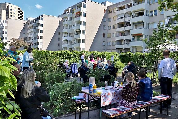 Menschen sitzen und stehen auf einer Grünfläche zwischen Hochhäusern; Kinder malen an einem Tisch.