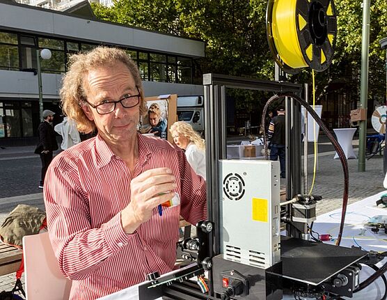 Herr Schellenberg vom Repair Café steht im Freien an einem Tisch mit einem 3D-Drucker und schaut in die Kamera.