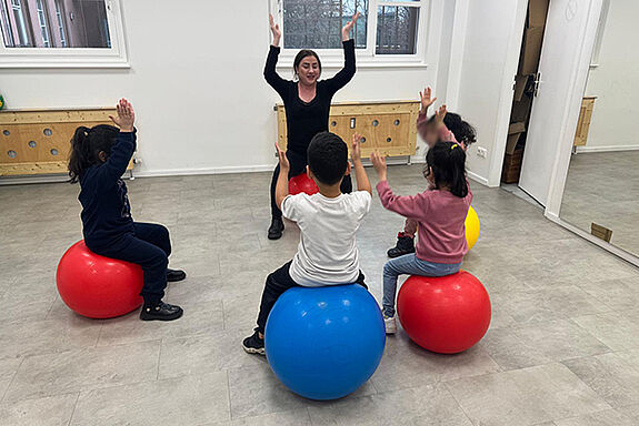In einem hellen Raum sitzt eine Gruppe Kinder auf großen Gymnastikbällen; eine anleitende Person steht vorn, hebt die Arme und die Kinder machen die Bewegung nach.