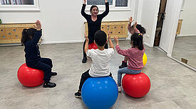 In einem hellen Raum sitzt eine Gruppe Kinder auf großen Gymnastikbällen; eine anleitende Person steht vorn, hebt die Arme und die Kinder machen die Bewegung nach.