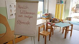 In einem hellen Raum einer Kindertagesstätte steht ein runder Tisch mit Bastelmaterialien sowie vier Stühlen. Davor steht ein Flip-Board mit dem Schriftzug Herzlich Willkommen zum heutigen Elterncafé. 