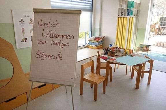In einem hellen Raum einer Kindertagesstätte steht ein runder Tisch mit Bastelmaterialien sowie vier Stühlen. Davor steht ein Flip-Board mit dem Schriftzug Herzlich Willkommen zum heutigen Elterncafé. 