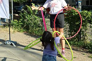 Ein kleines Mädchen spielt draußen mit mehreren farbigen Hula-Hoop-Reifen. Es ist von hinten zu sehen. Angeschnitten im Bild ein jugendlicher Mensch mit Hula-Hoop-Reifen und einem T-Shirt, auf dem „Kiezsportbus“ steht. Im Hintergrund ein begrünter Zaun und dahinter parkende Autos.