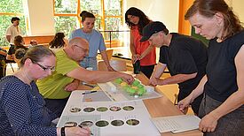 Mehrere Menschen diskutieren an einem Tisch anhand eines Platzmodells mit Bäumen und Gebäuden sowie mit Hilfe von Plakaten mit Fotos.
