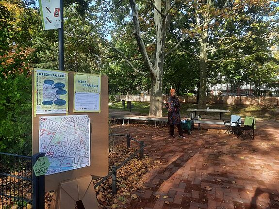 Eine Informationstafel mit Kiezplausch-Postern steht im Quartierspark, daneben eine Teilnehmerin, die sich in das Gesprächsformat einbringt.