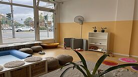 In einem hellen Raum der Kita Am Nettelbeckplatz befinden sich Sitzkissen, ein Bücherregal und Spielmaterialien. Eine große Fensterfront zeigt den Blick auf eine belebte Straße in Berlin-Wedding.