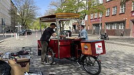 Ein umgebautes Lastenrad steht auf einer gepflasterten Straße. Zwei Männer stehen an dem Rad und unterhalten sich miteinander. 
