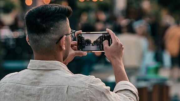 Ein Mann fotografiert mit seinem Smartphone eine belebte Straße bei Abendlicht.