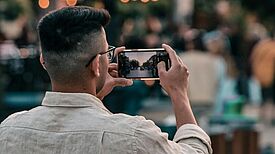 Ein Mann fotografiert mit seinem Smartphone eine belebte Straße bei Abendlicht.
