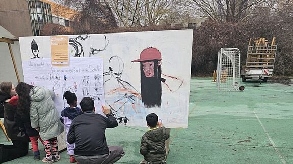 Mehrere Kinder stehen und knien auf einem Schulhof vor einer großen weißen Stellwand mit Zeichnungen und Schrift; ein Erwachsener zeigt auf die Wand, im Hintergrund stehen Sportgeräte und Bäume.