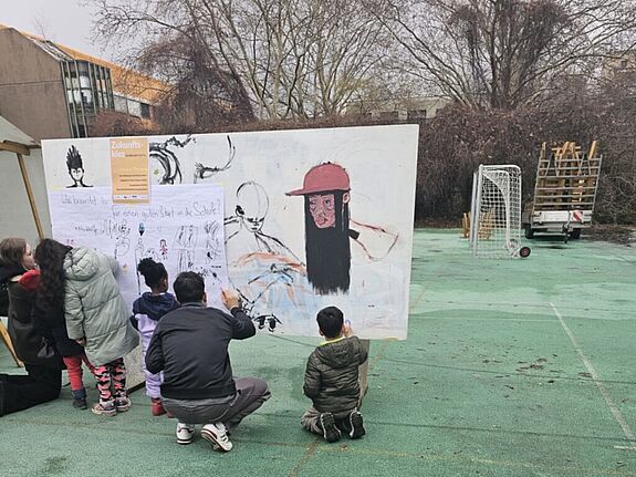 Mehrere Kinder stehen und knien auf einem Schulhof vor einer großen weißen Stellwand mit Zeichnungen und Schrift; ein Erwachsener zeigt auf die Wand, im Hintergrund stehen Sportgeräte und Bäume.