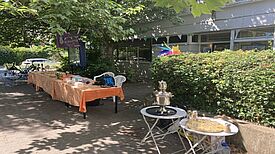 Ein langer Tisch mit Getränken und Tauschwaren steht im Schatten, der Samowar mit Gläsern auf Beistelltisch befindet sich beim Tauschmarkt daneben.