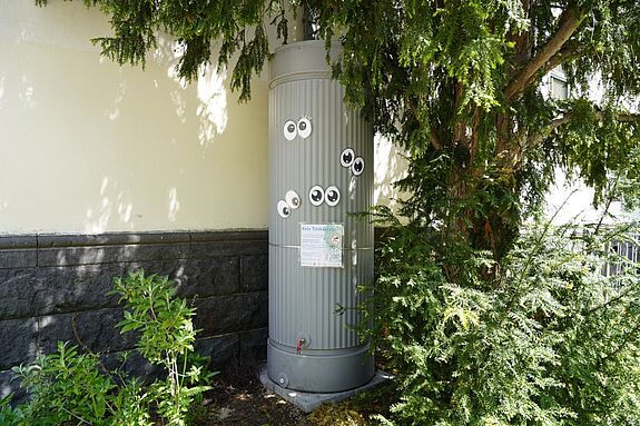 Regentonne an einer Hauswand mit Hinweistafel und aufgeklebten Augen.