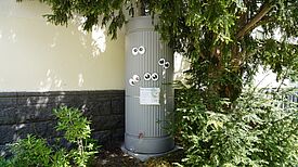 Regentonne an einer Hauswand mit Hinweistafel und aufgeklebten Augen.
