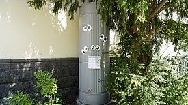 Regentonne an einer Hauswand mit Hinweistafel und aufgeklebten Augen.