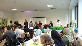 Während mehrere Personen vorne stehen, sitzt das Publikum beim Empfang für Engagierte in der Sonnen-Grundschule an festlich gedeckten Tischen.