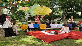 Kinder sitzen auf Bänken und schauen sich ein Puppentheater mit einem großen gelben Fisch im Freien an.