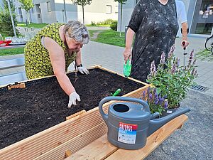 Zwei ältere Damen arbeiten an einem frisch angelegten Hochbeet mit angebauter Bank. Auf dieser stehen eine Gießkanne mit dem Schriftzug „Heißes Hellersdorf“ sowie Staudentöpfe. Im Hintergrund Neubauten, junge Bäume und grüne Wege sowie ein Tisch mit Gläsern und Wasserkaraffe.