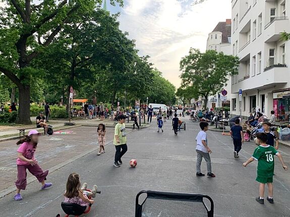Kinder spielen auf einer temporär gesperrten Straße im Quartier Pankstraße, während Erwachsene die lebendige Atmosphäre genießen.