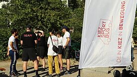Personen aus dem Team des KiezSportBus stehen im Freien zusammen und besprechen das Sportangebot vor einer Fahne mit der Aufschrift „KiezSportBus“.