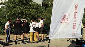 Personen aus dem Team des KiezSportBus stehen im Freien zusammen und besprechen das Sportangebot vor einer Fahne mit der Aufschrift „KiezSportBus“.