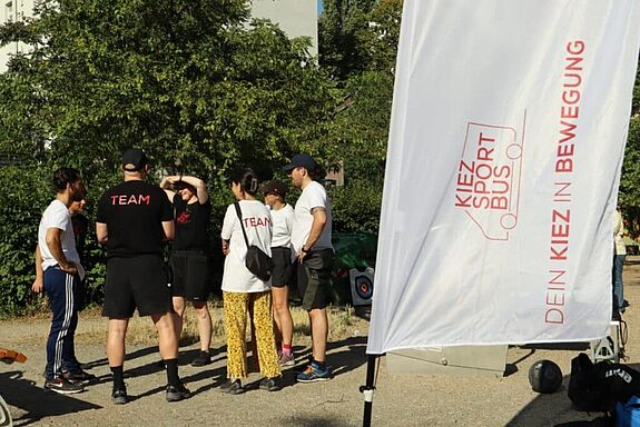 Personen aus dem Team des KiezSportBus stehen im Freien zusammen und besprechen das Sportangebot vor einer Fahne mit der Aufschrift „KiezSportBus“.