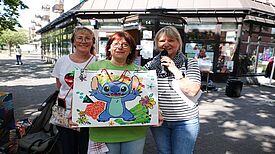 Drei Frauen stehen vor einem Gebäude, in den Händen halten Sie ; eine bunte Geschenktüte und ein Mikrofon.