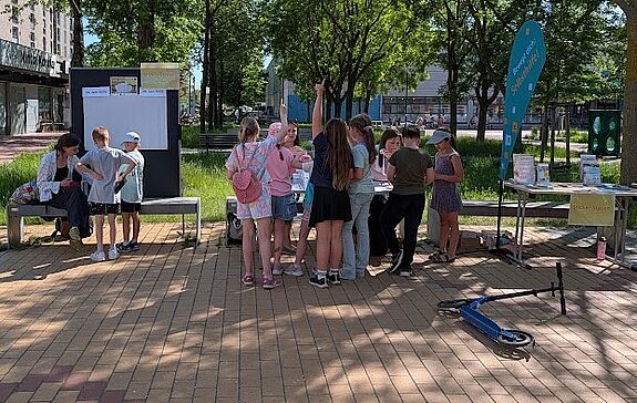 Eine Gruppe Jugendlicher steht an einem sonnigen Tag auf einem freien Platz vor Tischen mit dem Angebot „Back dir deinen Kiez“. 