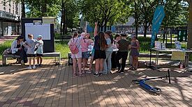 Eine Gruppe Jugendlicher steht an einem sonnigen Tag auf einem freien Platz vor Tischen mit dem Angebot „Back dir deinen Kiez“. 