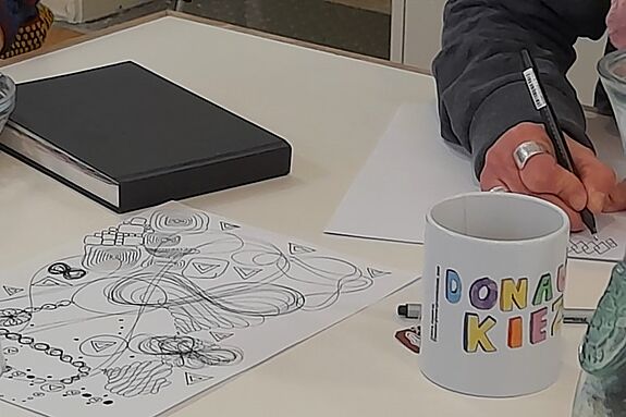 Eine Zeichnung, die bei dem Workshop entstanden ist, liegt auf dem Tisch, während eine Person etwas Neues zeichnet.